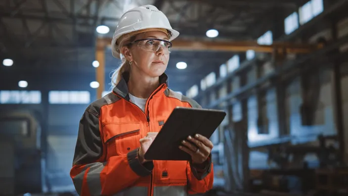 Professional Heavy Industry Engineer Worker Wearing Safety Uniform and Hard Hat Uses Tablet Computer. Serious Successful Female Industrial Specialist Walking in a Metal Manufacture Warehouse.