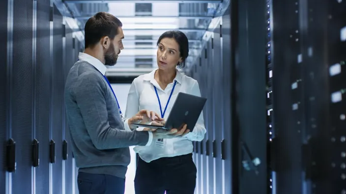 Male IT Specialist Holds Laptop and Discusses Work with Female Server Technician. They're Standing in Data Center, Rack Server Cabinet is Open.