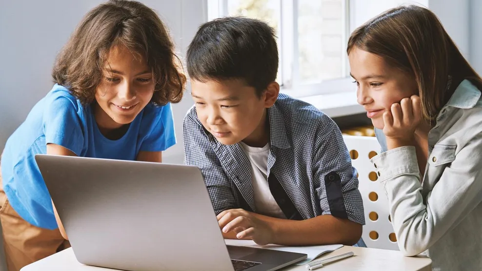 Diverse happy school kids using laptop computer together in classroom. Multicultural children junior students classmates learning online elementary education program class gathered at desk.