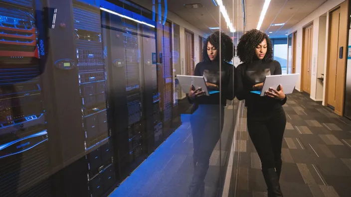 Young woman using laptop while working with server rack in contemporary data center hallway